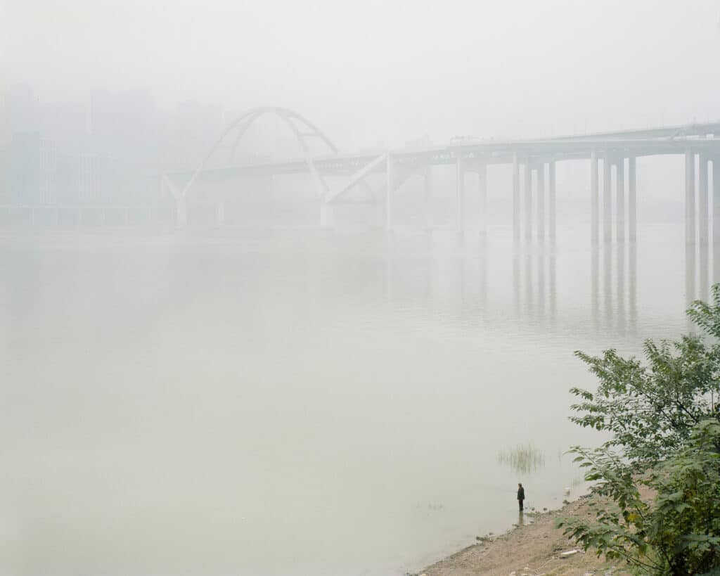 un puente sobre el agua visto a través de la niebla mientras un pescador está en la orilla