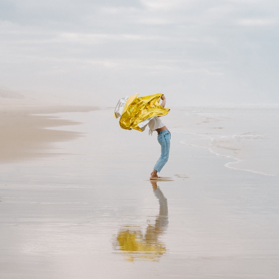 Frau in Jeans, T-Shirt und nackten Füßen steht im nassen Sand an einem bewölkten Strand und schüttelt leuchtend gelben Stoff. mit dem Titel "New summers" von Émilie Möri