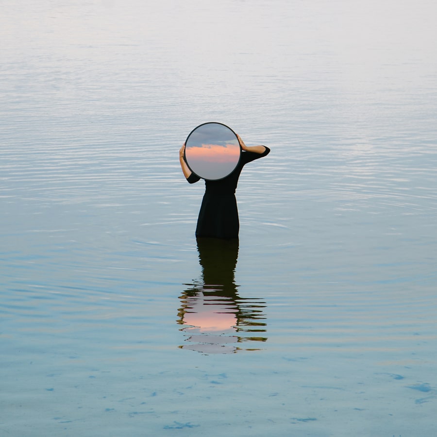 Eine Frau steht im Wasser, trägt ein schwarzes Kleid und hält einen runden Spiegel, in dem sich der Sonnenuntergang spiegelt. Mit dem Titel "Die Schatten meines Sees" von Émilie Möri