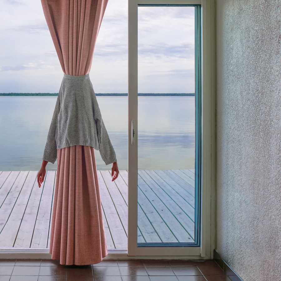 Blick aus einem Raum mit gläsernen Schiebetüren auf ein Deck über dem glasigen Wasser. Ein grauer Pullover liegt um einen langen rosa Vorhang, aus dessen Ärmeln die Hände einer Frau herausschauen. Bild von Émilie Möri