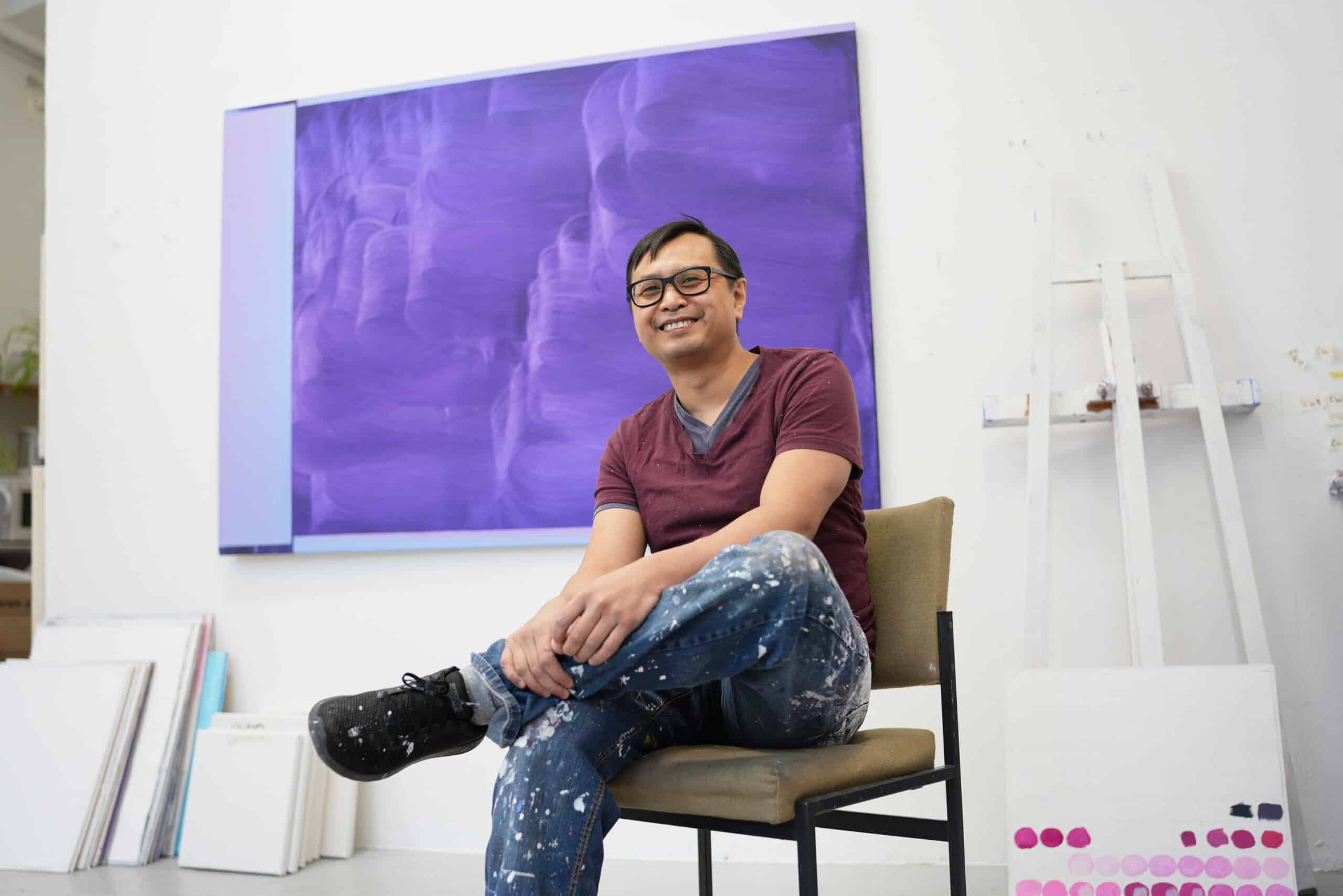 Retrato de Nam Duc Nguyen sorrindo e olhando para a câmera, sentado em uma cadeira em frente a uma pintura roxa com grandes marcas de pincel em seu estúdio.