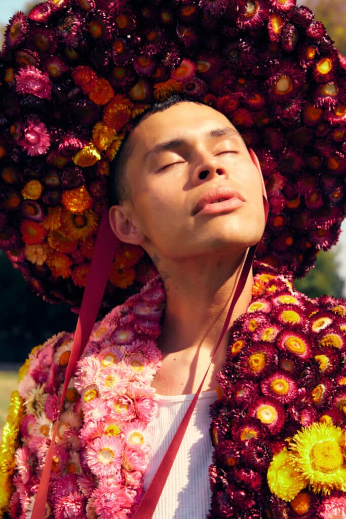 Man with his face turned toward the sun wearing a hat and suit covered in dried maroon, orange, pink, and yellow flowers