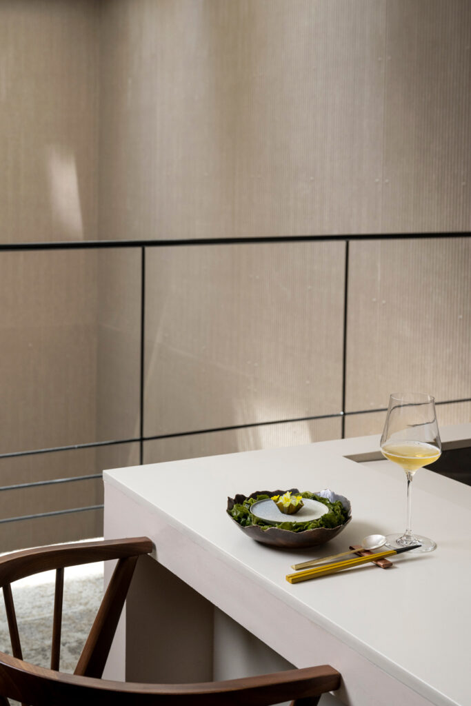Elegantes Gericht auf einem minimalistischen Tisch, neben einem Weinglas und Essstäbchen. Eine Wand mit neutraler Struktur ist der Hintergrund. Foto von Evan Sung