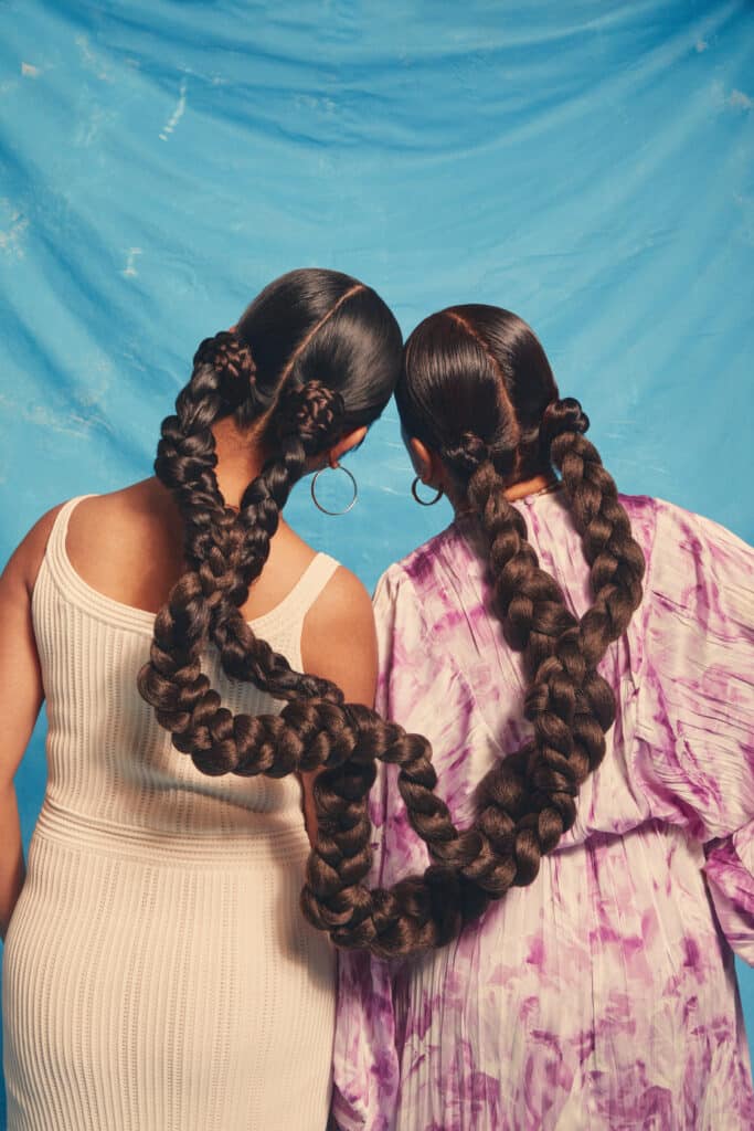Two woman with the backs to the camera, the tops of their heads resting together and their hair braided into two long connecting braids, arcing from one woman to the other.