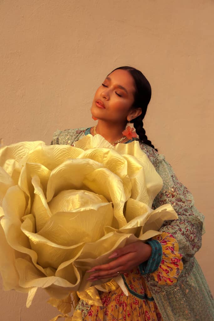 Mujer con la cara vuelta hacia su derecha mientras sostiene una flor de tela dorada más grande que la parte superior de su cuerpo.