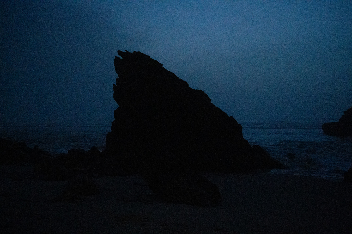 Una gran formación rocosa oscura se recorta contra un cielo y un océano azules y brumosos. La escena está tenuemente iluminada, lo que crea una atmósfera malhumorada y misteriosa. Fotografía de Marcia Simoes.