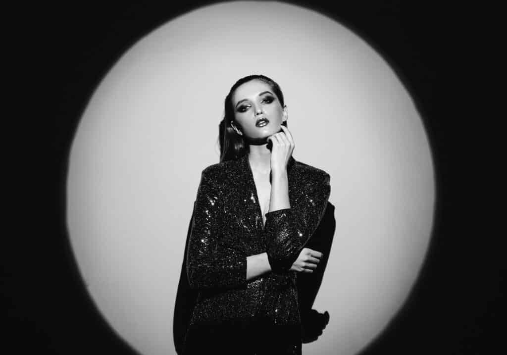 Black and white portrait of trendy girl with hair pulled back and stylish makeup in a shining dress standin g next to the wall in bright spotlight