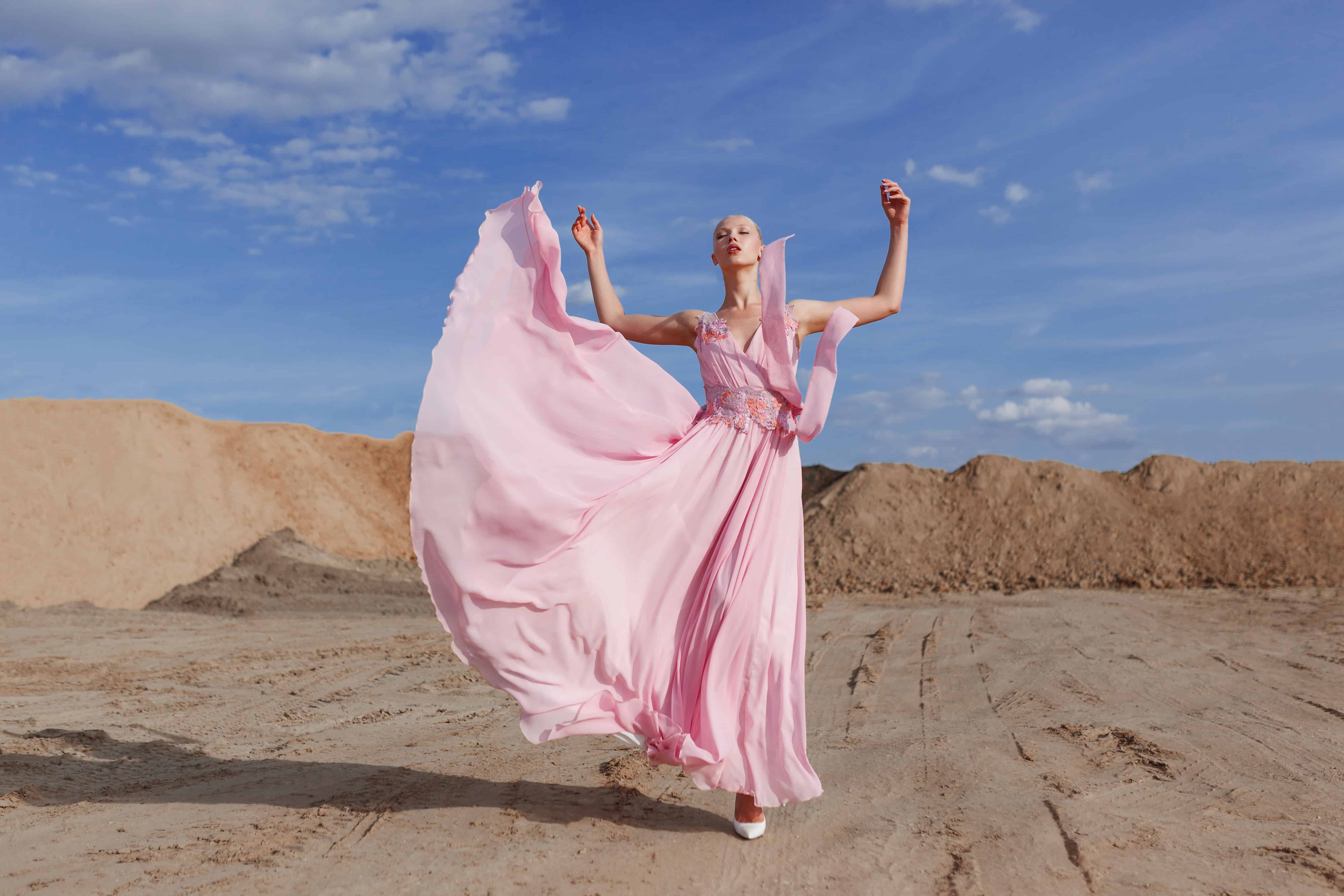 Full length portrait of a young beautiful blonde high girl in fashionable flying pink maxi dress poses in sand quarry on sunny summer day.