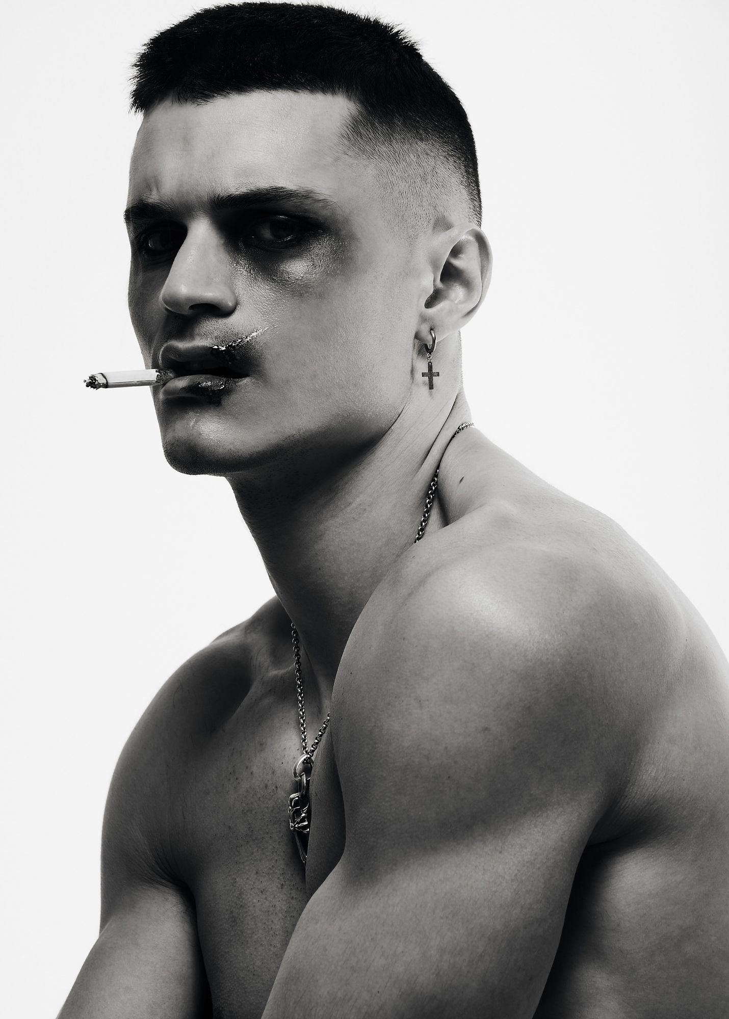 Black-and-white portrait of a shirtless man wearing a cross earring and smoking a cigarette