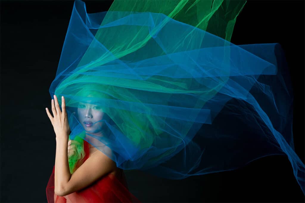 Woman wearing red and looking at camera through sheer blue and green fabrics in front of a black background. Photo by Edölia Stroud.
