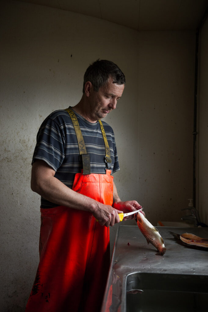 Ein Typ in orangefarbenen Overalls schneidet einen Fisch mit einem Messer an einer Edelstahltheke und Spüle. Foto von Evan Sung