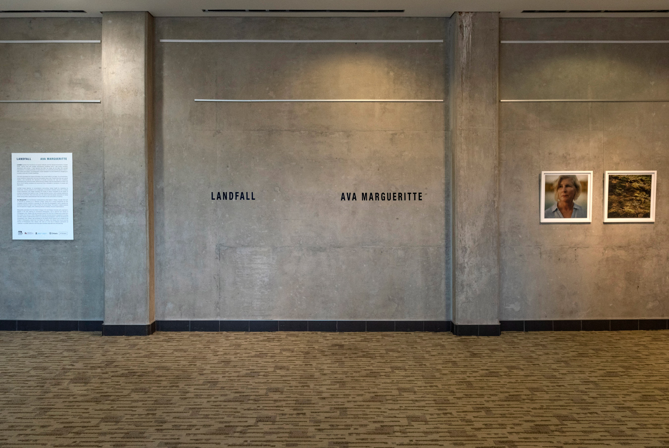 An art gallery wall displays the exhibition "Landfall" by Ava Marguerite, featuring a poster with exhibition details and two framed photographs: one a portrait of a woman and the other a landscape. The wall is concrete with black trim at the base, and the floor is carpeted.