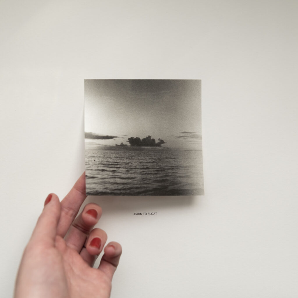A hand with red nail polish holds a square black and white photograph of a seascape with a cloudy sky, and the words "LEARN TO FLOAT" are printed below the image. The background is a plain white surface.