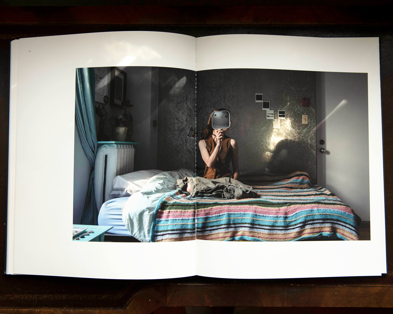 The image shows an open book with a photograph of a person sitting on a bed in a dimly lit room, holding a rectangular object in front of their face, obscuring it. The room features patterned wallpaper, a bed with a colorful striped blanket, and a door with sunlight streaming through.