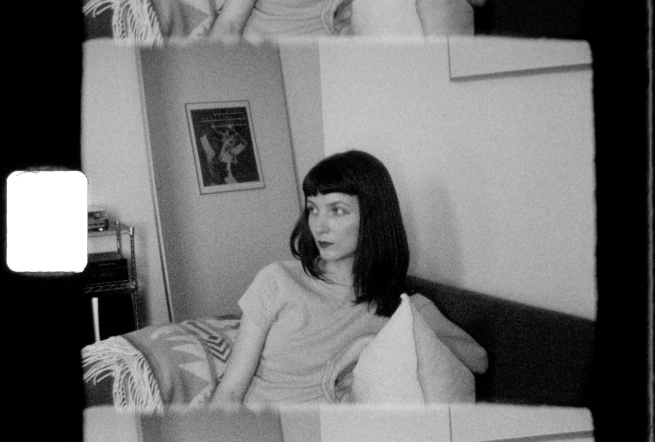 A black and white photograph shows a woman with dark, shoulder-length hair and bangs sitting on a couch, looking to the left. The image has a grainy texture and is a print from Super 8 film, with visible film borders and a bright, rectangular light leak on the left side.
