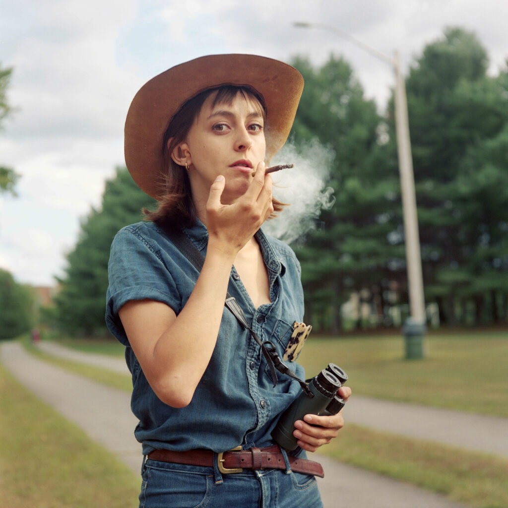 A person wearing a cowboy hat, denim shirt, and jeans stands on a path, smoking a cigarette and holding binoculars. The background includes trees and a cloudy sky.