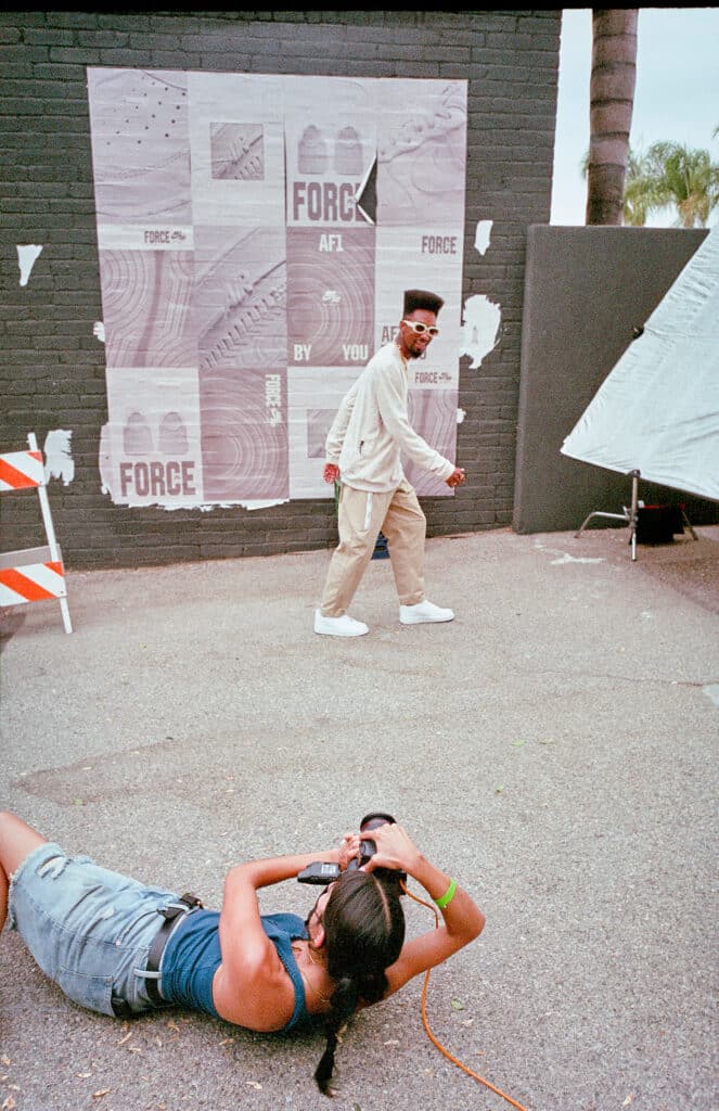 Brittany Bravo lying on the pavement with her camera for a different perspective while her subject poses in front of a wall covered in posters.