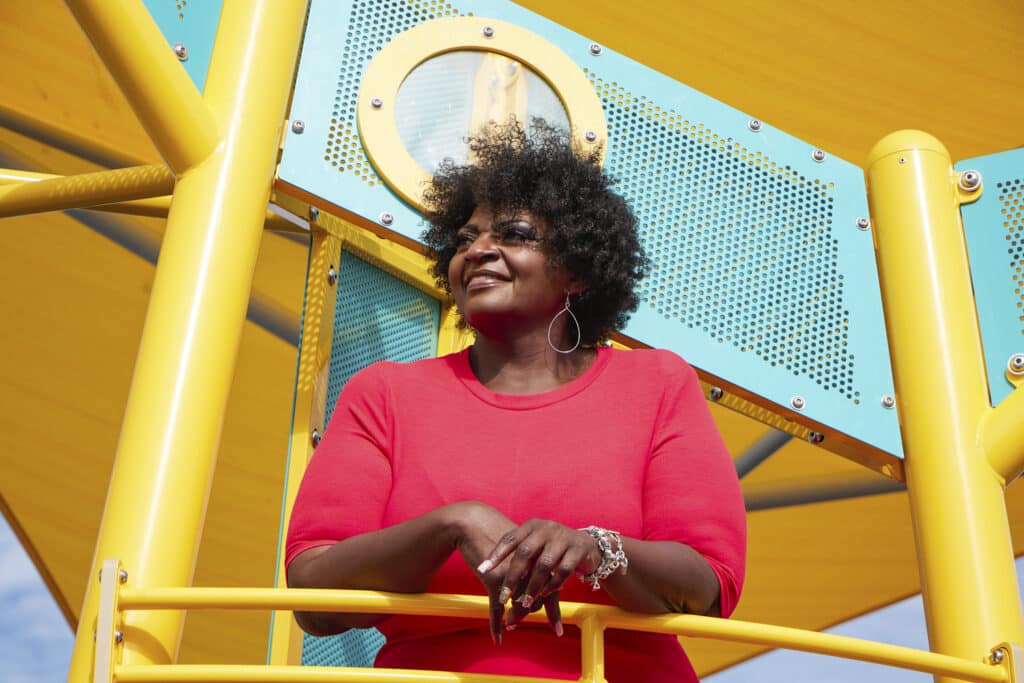 Mujer negra con top rojo de pie sobre una estructura de juego amarilla y aguamarina, mirando hacia el lado izquierdo de la imagen. Foto de J. Pamela Stills Photography