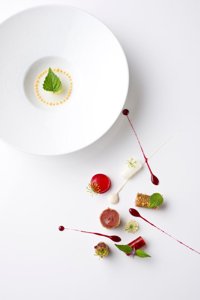 Gourmet-Gericht: Teller mit gelben Punkten und Kräutern, dazu kunstvoll angerichtetes Fleisch, rote und weiße Soßen, Beilagen auf weißem Untergrund. Foto von Evan Sung