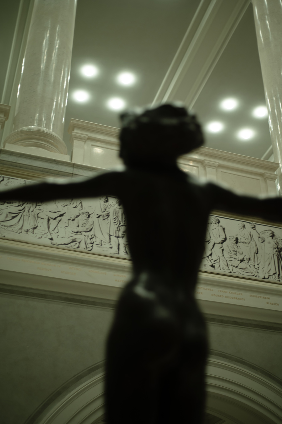 Una estatua borrosa y oscura de una persona con los brazos extendidos está delante de una pared con un bajorrelieve de figuras y texto, con columnas de mármol y un techo con luces empotradas al fondo. La estatua está desenfocada, mientras que los elementos del fondo están ligeramente más definidos. Fotografía de Marcia Simoes.