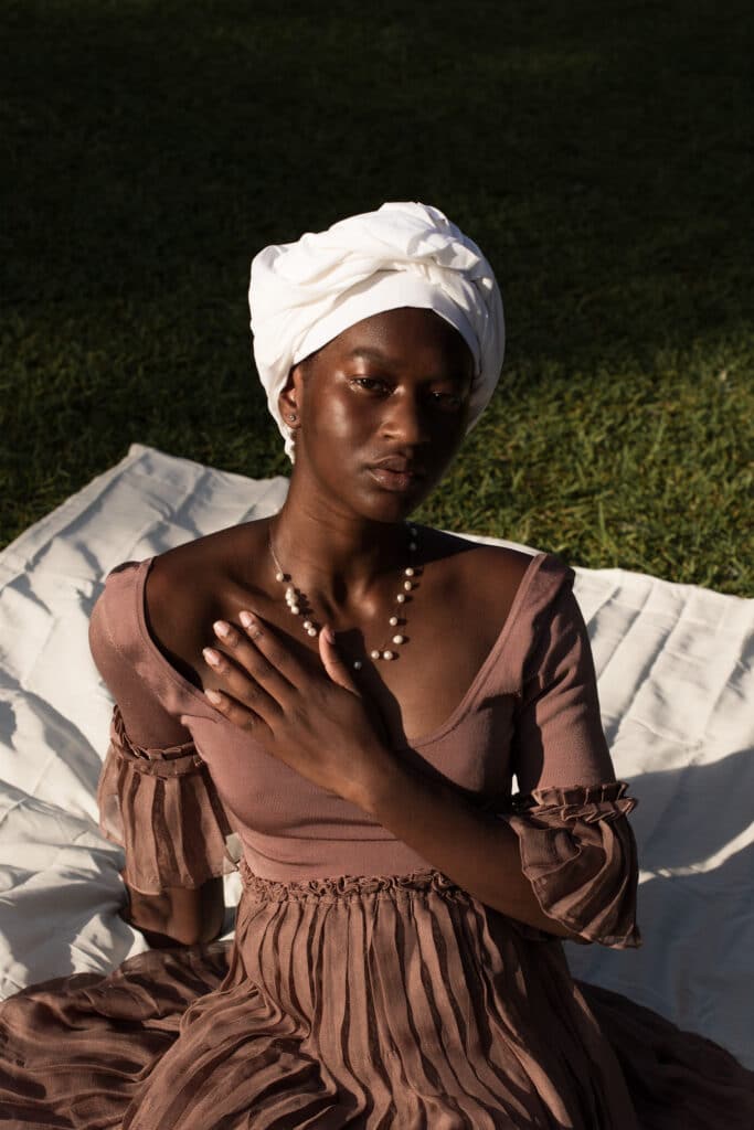 Black woman sitting on a white blanket on the grass, wearing a clay-colored dress and white turban, her left hand gently resting on her chest. Photo by Damola Akintunde.