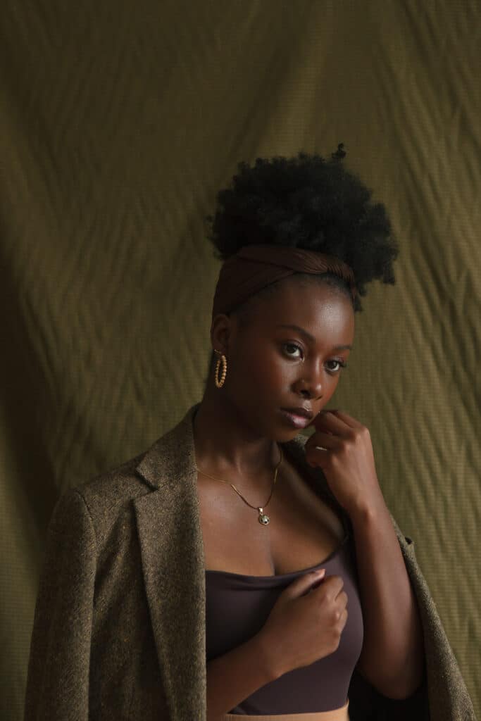black woman posing with one closed hand held softly by her cheek, wearing a mossy green tweed blazer and black top in front of a textured green backdrop. Photo by Damola Akintunde.