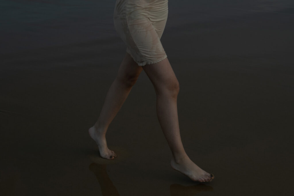 Se muestran las piernas de una persona caminando descalza sobre una superficie húmeda y oscura, posiblemente una playa, con la persona vestida con una prenda corta, de color claro y ribeteada con encajes. La imagen está débilmente iluminada, lo que crea una atmósfera sombría. Fotografía de Marcia Simoes.