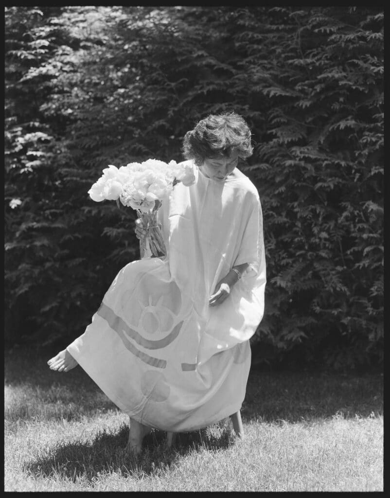 black and white image of a woman with short hair seated in a chair outdoors, wearing a voluminous white dress. Photo by Eno Inyangete.