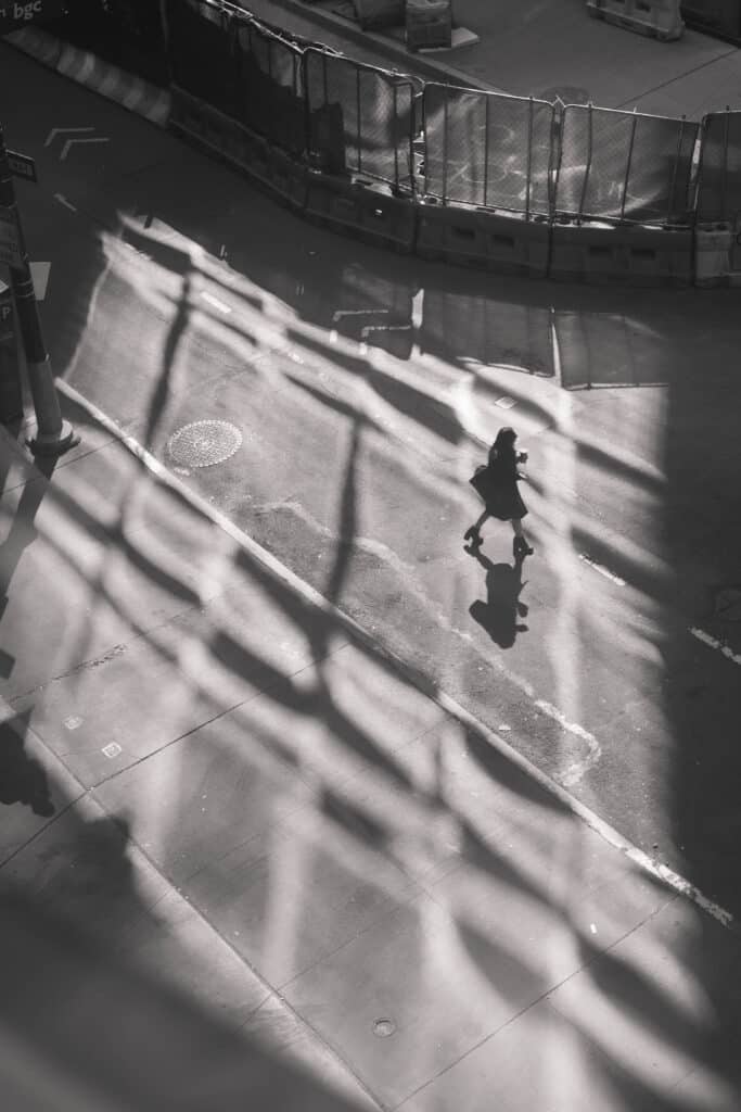 black and white image of a nearly empty street from above, light reflected from windows nearby and one figure crossing the street casting a shadow. Photo by Eno Inyangete.
