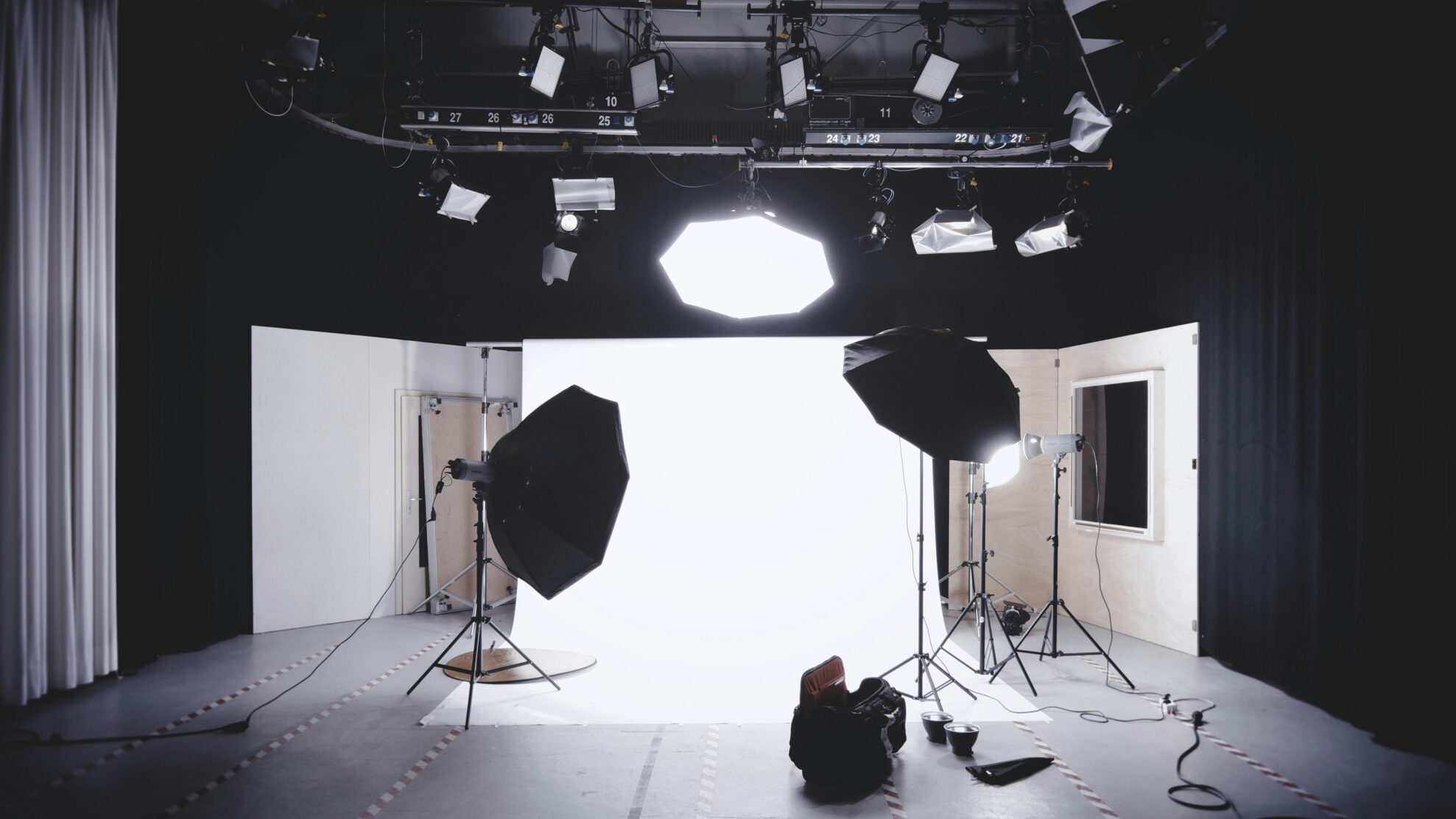 Ein voll ausgestattetes Fotostudio ist mit mehreren großen professionellen Beleuchtungskörpern, einschließlich Softboxen und Overhead-Stroboskopen, ausgestattet, die auf einen nahtlosen weißen Hintergrund gerichtet sind. Dieses Bild zeigt die umfangreichen Studioflächen, die Fotografen zur Verfügung stehen.