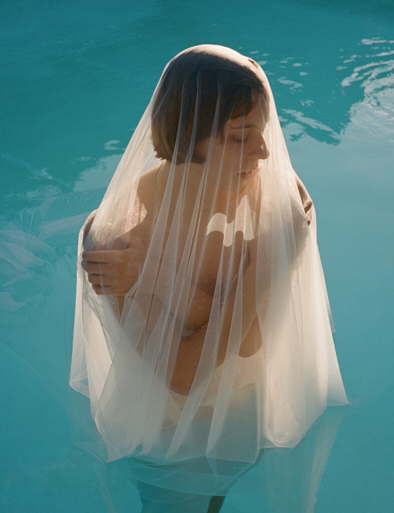 Mulher em uma piscina, meio submersa, coberta por um tecido transparente. Foto de Nick Prideaux