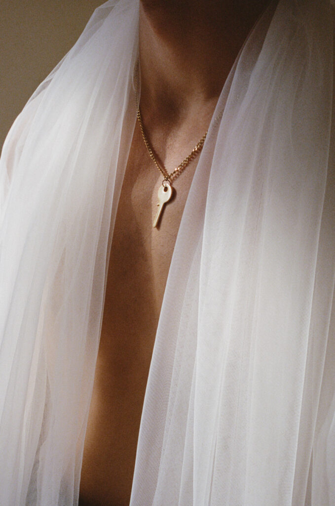 Close-up do pescoço e do tronco de uma pessoa envoltos em tecido branco transparente. Um colar de ouro com uma chave está pendurado no pescoço. Foto de Nick Prideaux.