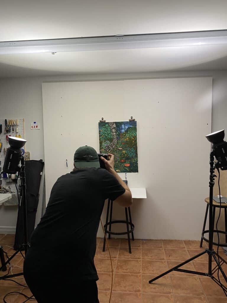 œuvre d'art colorée et détaillée de Stanzie Tooth accrochée au mur blanc pendant qu'Ibrahim Abusitta prend des photos pour documenter l'œuvre.