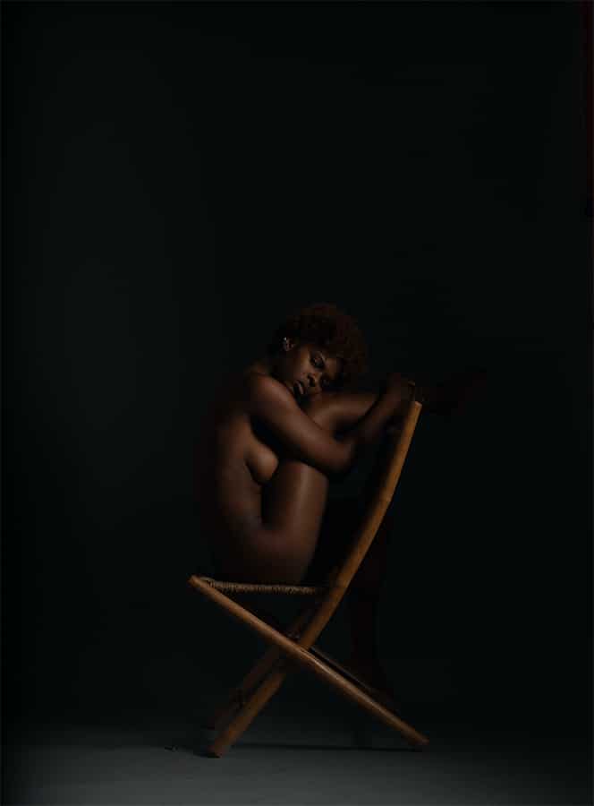 nude black woman sitting backward in a bamboo chair, her legs resting on the back and her arms wrapped around them, in front of a black background. Photo by Jessica Bethel.