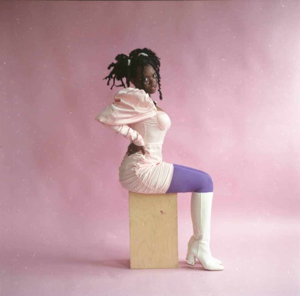 Black woman with braids wearing a light pink dress, purple tights and knee-high white boots, seated on a wooden cube in front of a pink backdrop. Photo by Jessica Bethel.