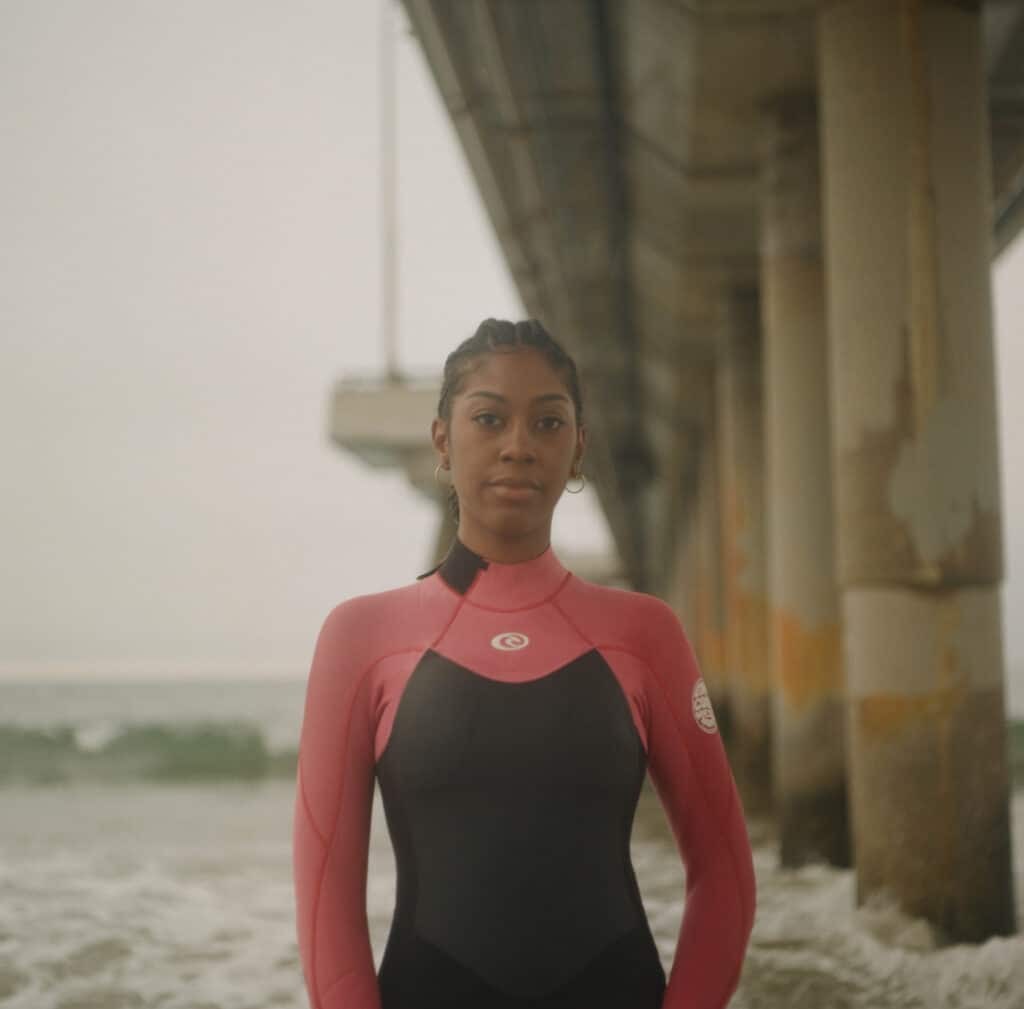 Garota surfista negra vestindo uma roupa de mergulho rosa e preta, perto de ondas quebrando e de um calçadão de concreto no oceano. Foto de Jessica Bethel.