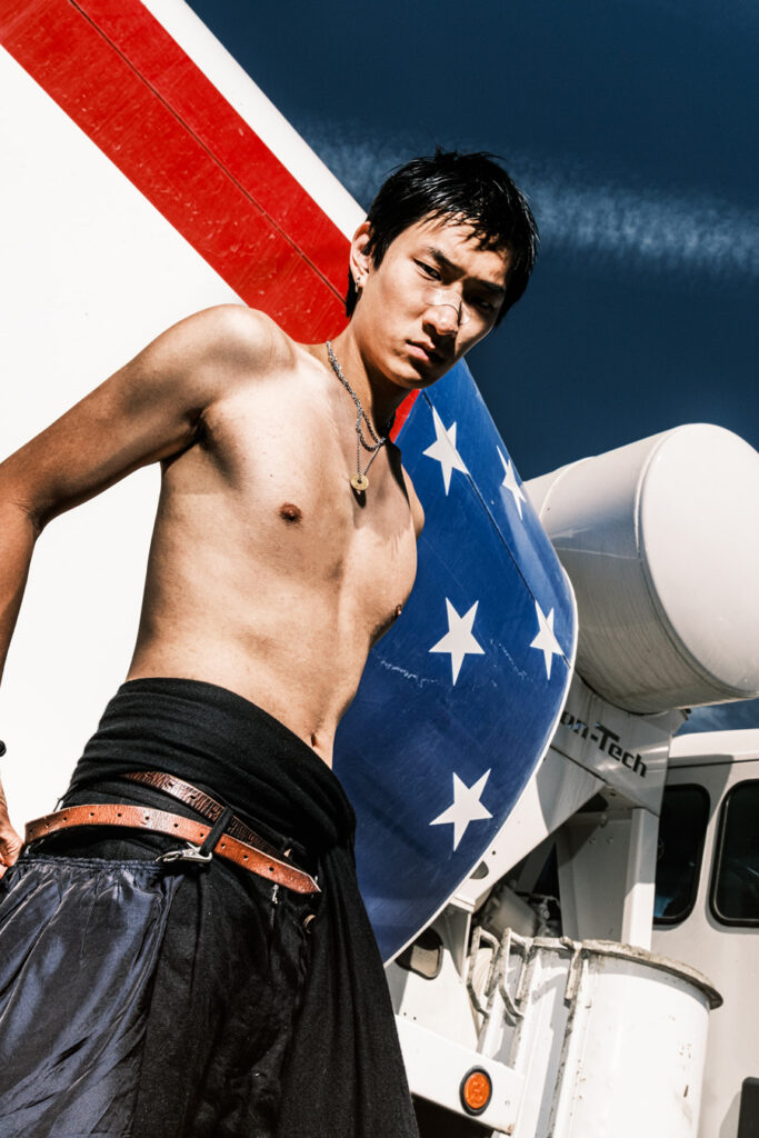 Ein asiatischer Mann ohne Hemd und mit einem Verband auf der Nase steht vor einem weißen Truck mit einer rot-weiß-blauen amerikanischen Flagge. Er trägt ein schwarzes Tuch um seine Taille und einen braunen Gürtel. Foto von Joshua Conliffe.