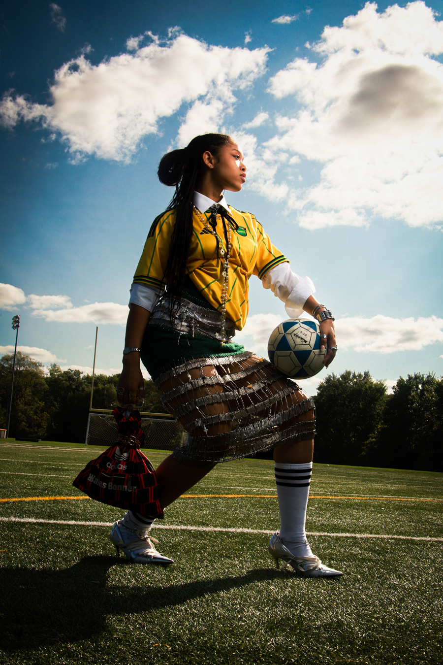 Eine Person mit langen dunklen Haaren steht auf einem Fußballfeld und trägt ein gelb-grünes Fußballtrikot, einen Rock aus Sicherheitsnadeln, weiße Socken mit schwarzen Streifen und silberne Absätze. Sie hält einen Fußball auf dem Oberschenkel und eine rot-schwarze Tasche in der Hand. Im Hintergrund sind ein blauer Himmel und Wolken zu sehen. Foto von Joshua Conliffe.