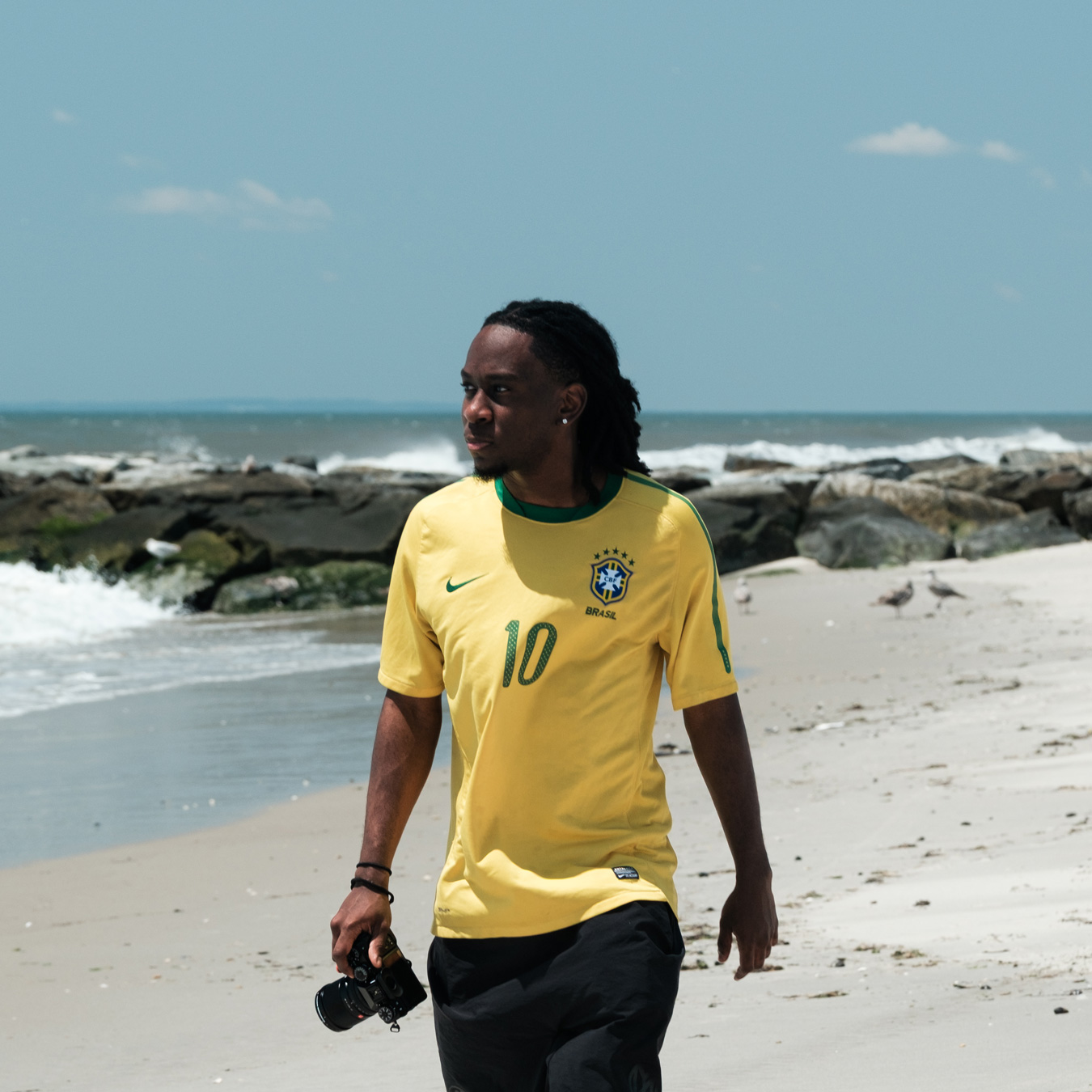 Ein Mann mit Dreadlocks, der ein gelbes brasilianisches Fußballtrikot und schwarze Shorts trägt, läuft an einem Sandstrand entlang und hält eine Kamera in seiner rechten Hand. Hinter ihm schlagen Wellen gegen einen felsigen Wellenbrecher unter einem hellen Himmel. Porträt des Fotografen Joshua Conliffe.