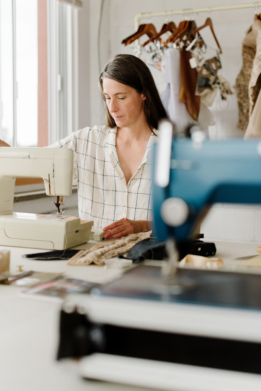 Eine Frau in einem karierten Hemd näht konzentriert an einer alten Nähmaschine in einem hellen Atelier, während im Hintergrund andere Nähmaschinen und Kleidungsstücke zu sehen sind. Foto von Sarah Bodri.
