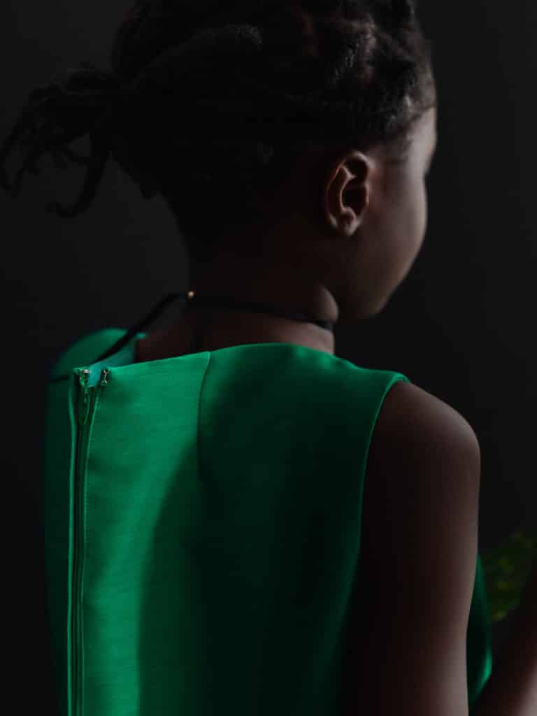 Une jeune fille noire, dos à l'appareil photo, dans une robe verte surdimensionnée, la fermeture à glissière dépassant raidement derrière elle. Photo prise par Karene-Isabelle Jean-Baptiste.