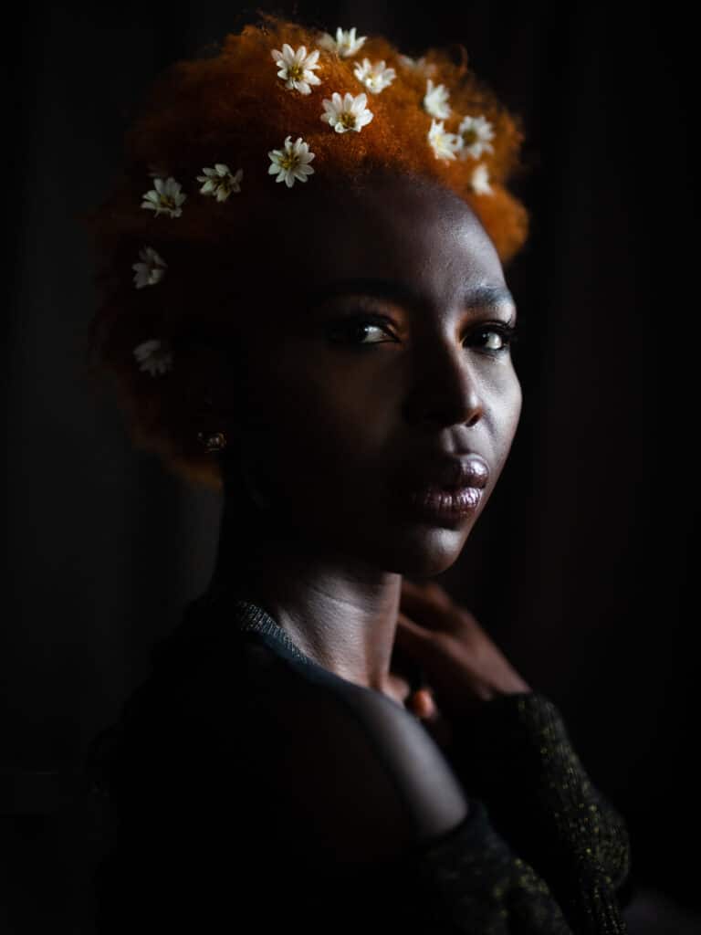 Foto de Karene IsabelleJean Baptiste. Foto de rosto de uma mulher negra sob luz dramática, com seu cabelo curto de textura natural tingido de laranja e pequenas margaridas colocadas artisticamente em seu cabelo.