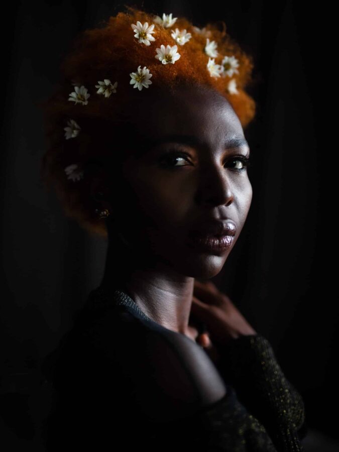 Photo by Karene IsabelleJean Baptiste. Headshot of black woman in dramatic light with her short, natural texture hair dyed orange and small daisies placed artistically in her hair.