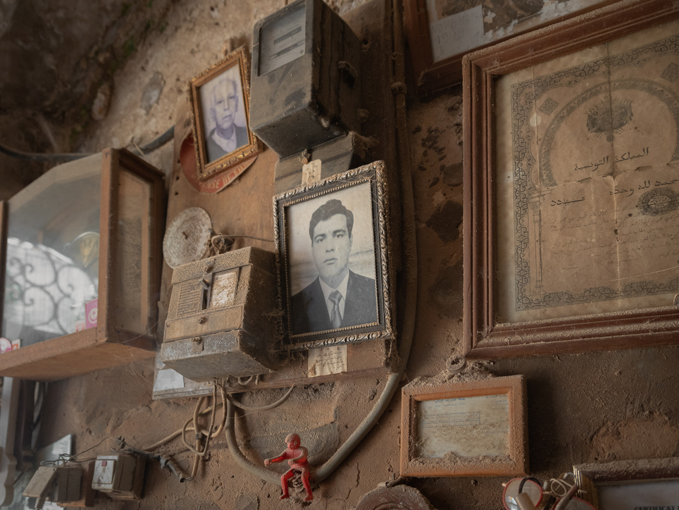 A dusty wall is adorned with framed photographs, certificates in Arabic, and old electrical meters, creating a vintage and somewhat neglected display. A small red toy figure clings to the wall amidst the collection. Photo by Skander Khlif.