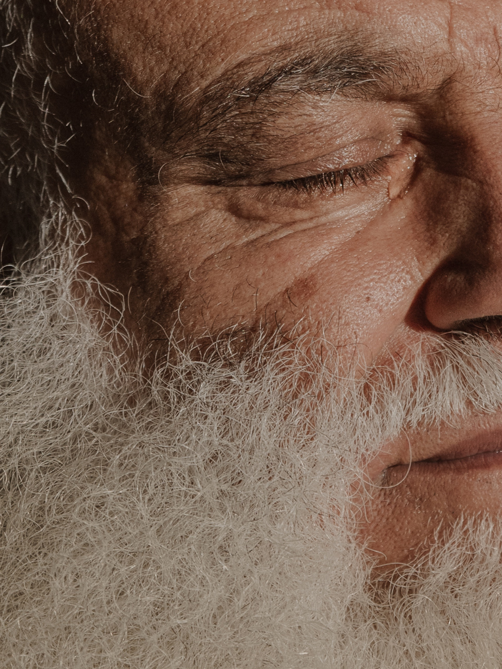 A close-up shot shows the right side of an older person's face, featuring a closed eye with wrinkles, a prominent nose, and a portion of their mouth, all partially obscured by a thick, white beard. The skin has visible texture and details, illuminated by a warm light. Photo by Skander Khlif.