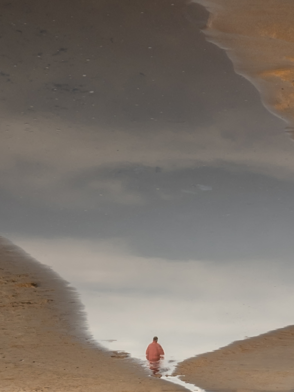 The image shows the reflection of a beach scene in a puddle, with a person in a red shirt standing in the water. The sky and clouds are reflected in the puddle, creating a surreal and inverted perspective. Photo by Skander Khlif.