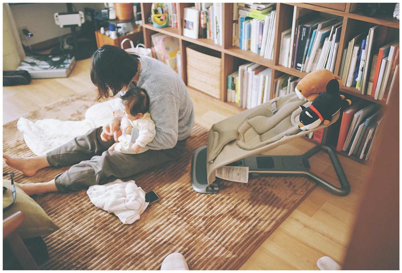 A person sits on a brown rug, holding a baby. A baby bouncer with stuffed animals sits nearby. A bookshelf filled with books is in the background. Photo by Ko Okamoto.