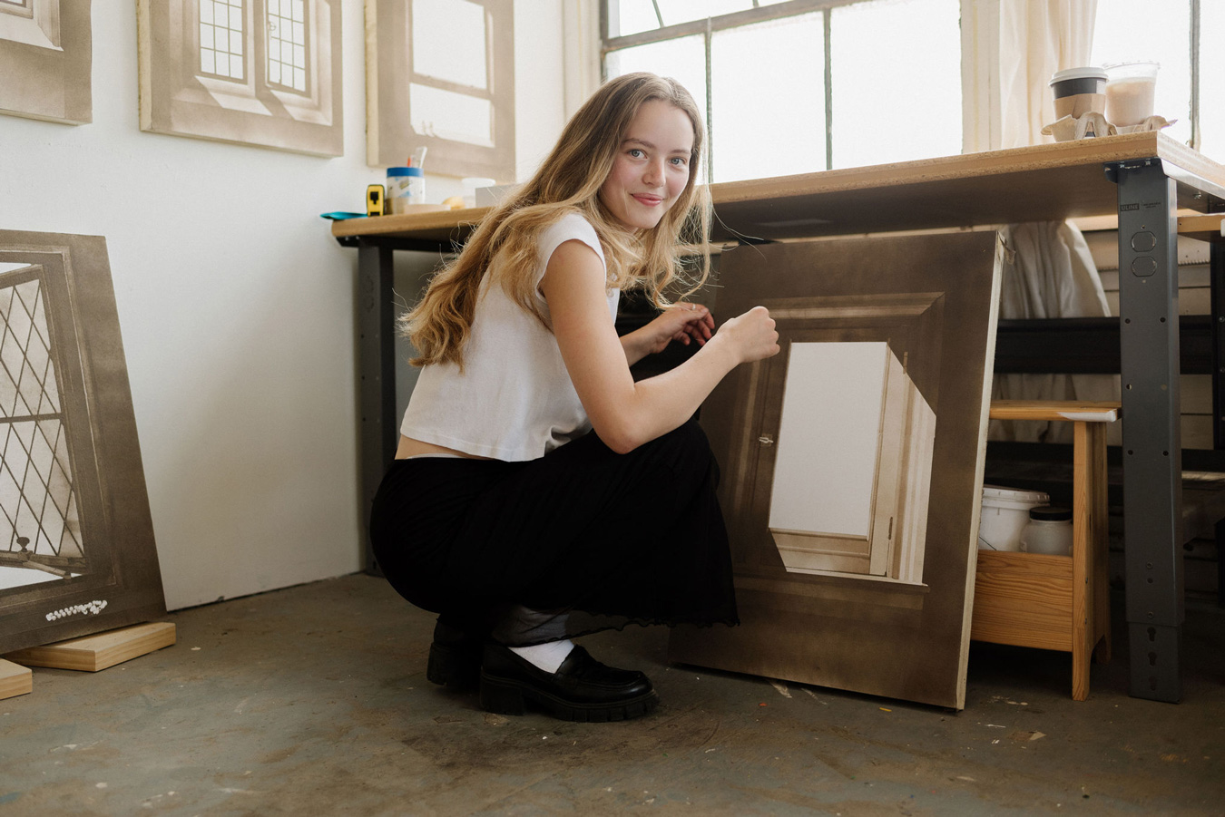 Eine junge Frau mit langen blonden Haaren hockt in einem Kunstatelier und lächelt in die Kamera, während sie von ihren Kunstwerken umgeben ist, die Fenster darstellen. Sie trägt ein weißes T-Shirt, einen schwarzen Rock und schwarze Halbschuhe mit weißen Socken. Foto von Sarah Bodri.