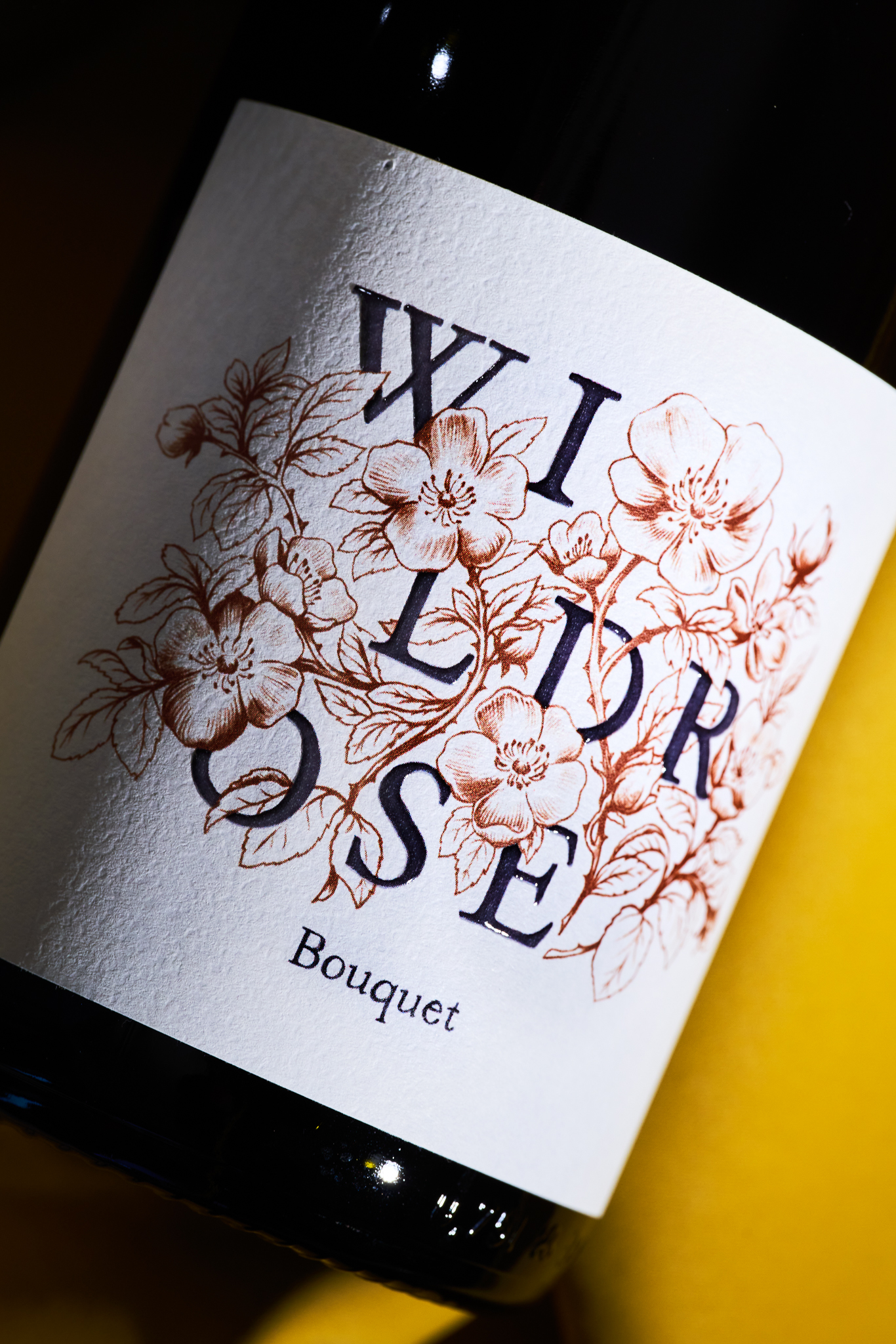 A close-up shot of a wine bottle label featuring the words Wild Rose in a stylized dark blue font intertwined with brown line drawings of blooming wild roses and leaves, with the word Bouquet printed in smaller text below.