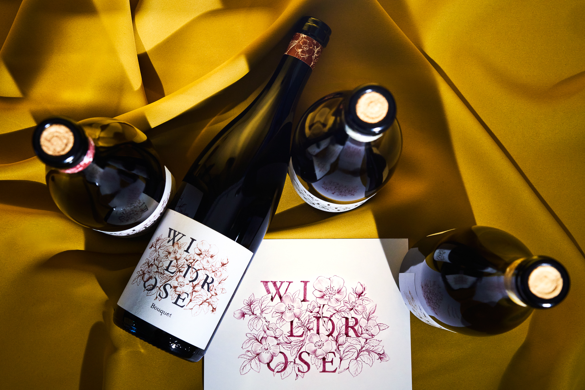 Four dark glass wine bottles labeled Wild Rose Bouquet are arranged on a draped mustard yellow fabric alongside a matching floral card.